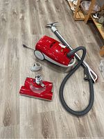 Red Elegance TrueHEPA plug-in vacuum cleaner with hose, metal wand, carpet extension and two additional attachments, shown on wood floor