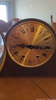 Front view of clock showing gold-tone dial, Roman numerals, and open glass bezel