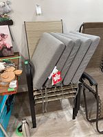 Four stacked patio chairs with dark metal frames and tan woven seating surfaces. Four grey cushions stacked on top with tie strings visible, tags attached.