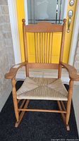 Full front view of wood framed rocking chair with curved armrests, vertical spindle backrest, and woven wicker seat in front of yellow door.