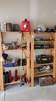 Wide view of two pine shelving units filled with assorted tools, containers, boots, and toolboxes in garage.
