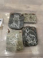 Four soapstone rectangular plaques with white engraved images including an owl, bear, person in traditional clothing, and a stylized animal, all with leather cords.
