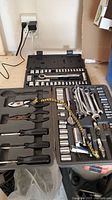 Two open tool cases filled with ratchets, sockets, wrenches, and pliers laid out on a table against a wall socket.