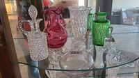 Shelf displaying all items in the lot: cranberry glass vase, green glass vase, clear crystal vase, glass oil decanter, crystal bell, and glass paperweight.