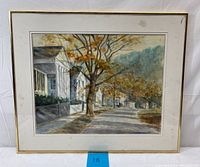 Framed watercolor painting showing street scene with white houses, trees with autumn leaves, and a distant figure on sidewalk, natural light, minor frame wear.