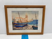 Framed watercolor painting showing a coastal beachscape with boats and buildings, the horizon line near the upper third of the scene.