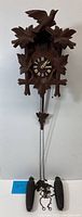 Front view of vintage cuckoo clock showing carved bird atop, wooden leaves, Roman numeral dial, and two pine cone weights attached with chains.