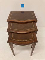 Three stacked vintage nesting tables showing wavy wooden edges and brown leather tops with gold borders.