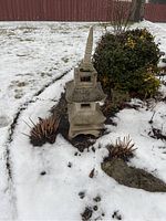 Front angle view of weathered cement pagoda statue in snowy garden setting with some shrubbery around it.