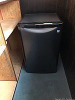 Photo of Danby black bar fridge closed in a tight corner space on carpeted floor.