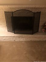 Black metal fireplace screen with curved top and mesh panels placed in front of a fireplace opening as seen from a distance.