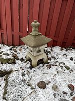 Front view of the cement garden pagoda placed outside on snow-covered ground with rocks around it and a red fence in the background.