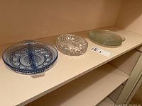 Four glass items on a shelf - blue divided dish, clear round ashtray, and two other clear glass plates