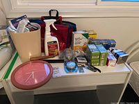 Wide view of the household items including the pink wall clock, cleaning bottles, waste basket, light bulb boxes, and various small tools and accessories arranged on a white table.