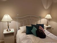 Photo shows a bedroom setting with the two matching metal swing-arm table lamps on white nightstands beside a white metal bed frame with pillows.