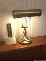 Photo showing the brass desk lamp lit, standing next to the Nikko Seiki metronome on a wooden surface with wallpaper background.