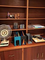 Overview of items on a wooden shelf including fan, file folders in holder, CDs in spindle, hole puncher, paper cutter, clipboard, and mouse pad