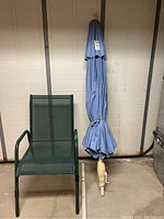 Patio chair with arms and mesh seat/back next to folded blue patio umbrella with white pole.