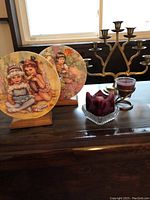Two decorative Wedgwood My Memories series plates on wooden stands with a five-armed candlestick and a glass candle holder with a red candle on a wooden surface.