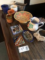 Collection of pots, straw basket, two colorful ceramic tiles, a white floral pitcher and stoneware vessel on wooden surface