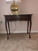 Front view of dark wooden side table with cabriole legs and brass handle drawer under a lamp.