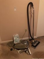 Full view of the vacuum cleaner with hose, metal wand, power head, and vacuum bags to the left. Carpeted floor, beige walls.