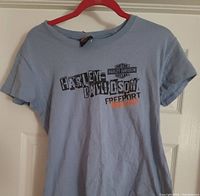 Light blue women's T-shirt hanging on red hanger, showing full front view with Harley Davidson Freeport Bahamas print