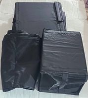 Three black fabric wardrobe boxes and garment bags folded flat, showing the texture and material