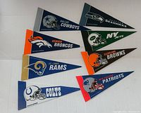 17 vintage NFL mini pennants laid out in two groups on a white surface. Visible teams include Cowboys, Seahawks, Broncos, Rams, Colts, Patriots, Jets, Browns, Steelers, Packers, Bears, Saints, Ravens, Panthers, Raiders, Falcons, and Cardinals.