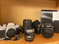 Minolta film camera body, two manual focus lenses, Panasonic Lumix camera, and carrying case on wooden shelf