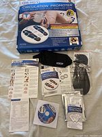 Photo of the box, device with attached foot stimulation pads, black pouch, instruction manual, replacement TENS pads, CD, and other papers laid flat.
