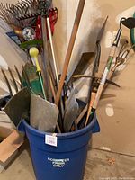 Large blue garbage bin filled with multiple garden tools including rakes, shovels, hoes, and forks.