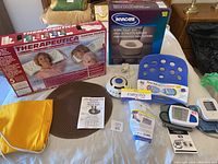 Full view of health and safety items on bed including pillows box, toilet seat box, posture seat, blood pressure monitors, health mats, white noise machine