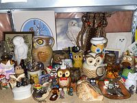 Wide view of large assortment of owl decor items on shelves and table, showing ceramic pitcher, white carved owl, needlepoint, figurines, and artworks.