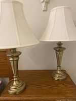 Pair of brass table lamps with white fabric shades on a wooden surface against wall.