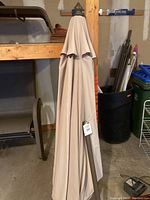 Closed beige patio umbrella with finial top and tag attached, standing in a basement or storage area.