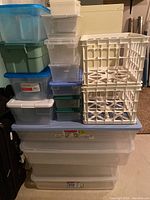 Photo showing a stack of various plastic storage containers including large clear bins at the bottom, smaller translucent colored boxes, and two white plastic milk crates.
