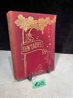 Photo of the front cover of the antique book The Dew Babies showing red hardcover with embossed gold decoration and figures