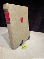 Photo of the book's front cover showing beige textured hardcover with an embossed red emblem and red and black text on the spine.