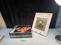 Lot of 5 coffee table books displayed on a table; one upright 'Treasures of the Louvre' book and four stacked horizontally including 'Brera Milan' among others.