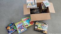 Photo showing a cardboard box with multiple kids' board games including Mastermind, Minions Trouble, and Discovery Kids Super Slime Lab, with more kits around the box.