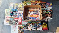Flat lay of various vintage sports yearbooks, scorebooks, and magazines spread around a cardboard box filled with sports bumper stickers.