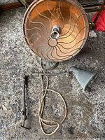 Top view of large circular metal reflector of antique industrial heater with base and aged fabric-wrapped electrical cord with plug, and articulated metal arms with cone shaped lamp shade on the ground