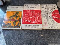Three vintage paperback books placed on a black countertop: 'Canadian Art: Its Origin and Development' by William Colgate on the left, 'Antiques in Ontario 1969-70' in the center, and 'Antiques in Ontario 1965-66' on the right.