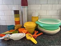 Wide view of vintage Tupperware storage containers, measuring spoons, and utensils, metal juice glasses stacked with red plastic cups on top, and a yellow Thermos bottle.