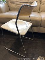 White plastic Emu chair with chrome tubular frame shown from side angle on dark wood floor in front of beige couch.
