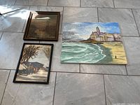 Three framed paintings placed on tiled floor including seaside, winter sleigh, and sepia scene