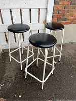 Set of three retro bar stools with white metal frames and black round seats shown from a side angle.