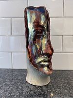 Front left side view of the pottery face sculpture showing textured base and iridescent glaze.