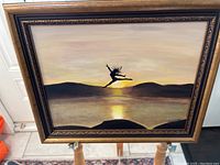 Full view of framed vintage painting depicting a dancer's silhouette leaping at sunset over water.